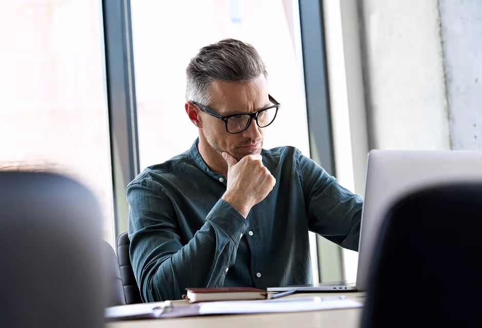Thoughtful man in glasses at a laptop in an office, hand on his chin