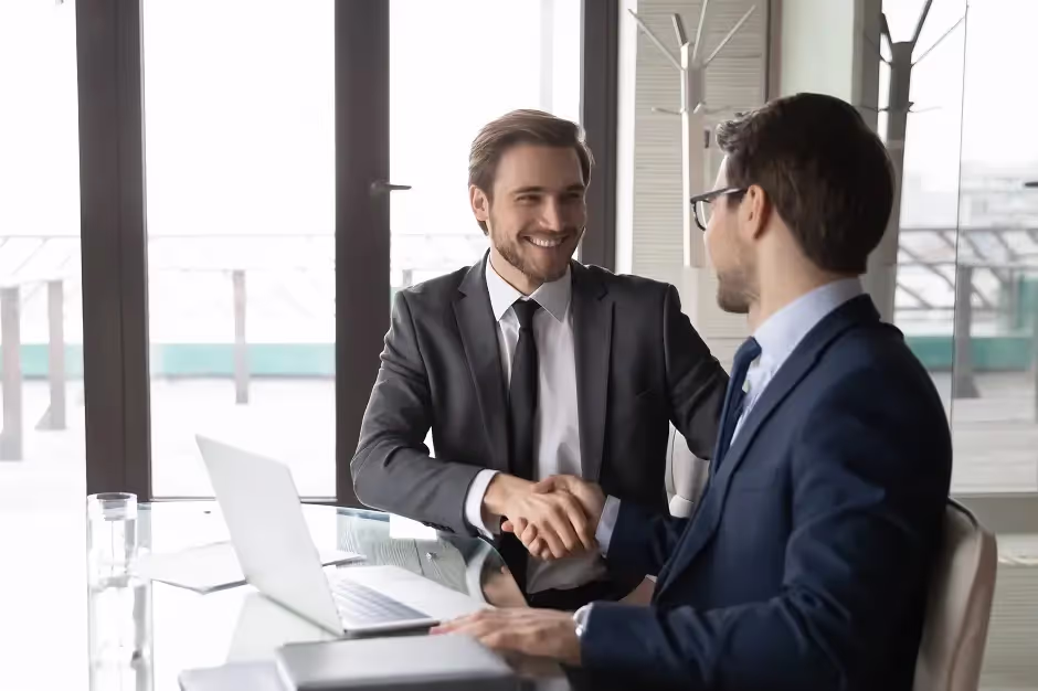 Two businessmen in suits smiling and shaking hands at an office desk