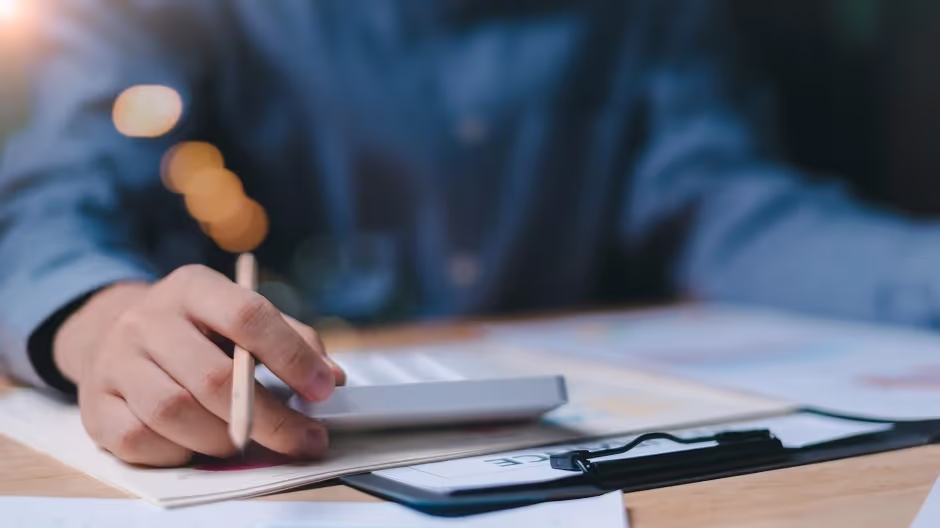 Hand holding a pencil over paperwork and a calculator on a desk