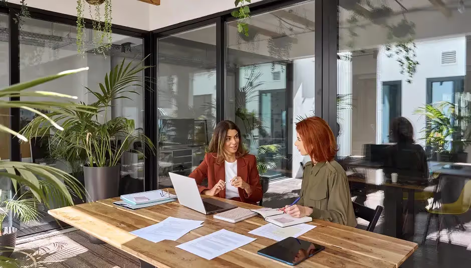 Two women discussing documents at a table in a bright office with plants
