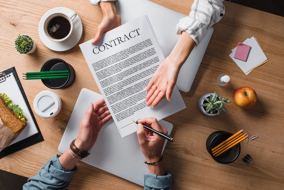 Two people at a wooden desk, one handing a contract to the other to sign