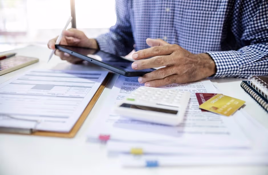 Businessman using a tablet beside invoices, a calculator and credit cards on a desk