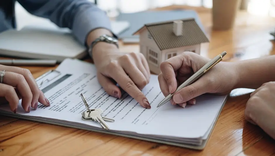 Two people signing a document with house keys and a small model house on the table