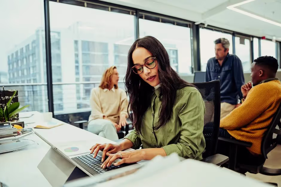 Woman in glasses typing on a laptop in a busy open-plan office