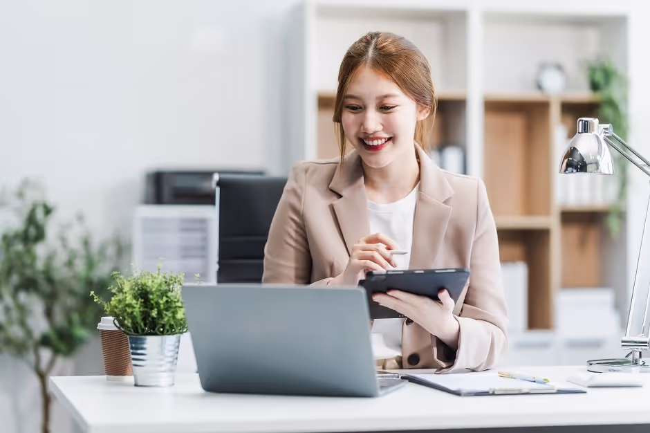 Businesswoman smiling as she works on a tablet and laptop at her desk