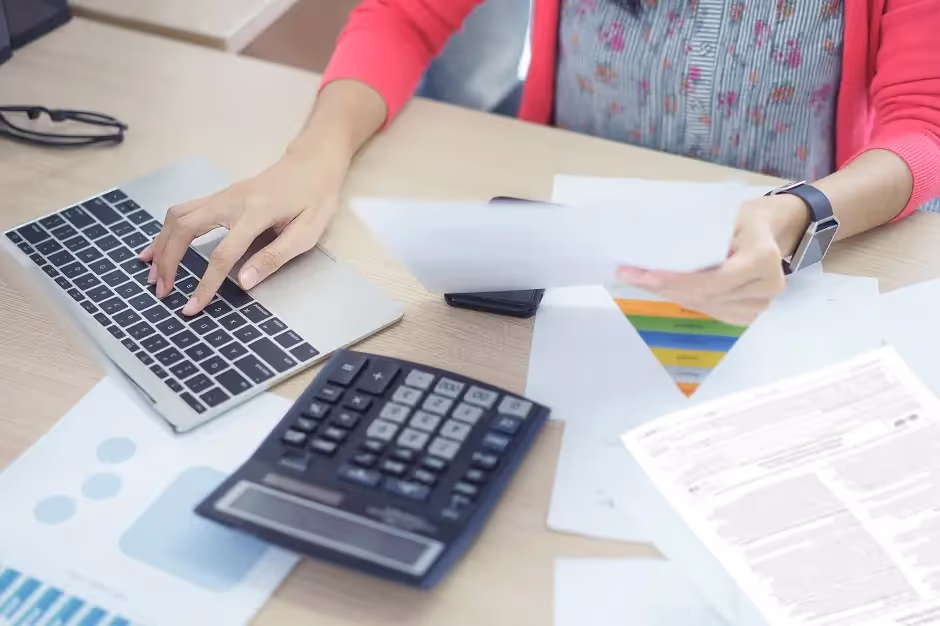 Person working on a laptop beside a calculator and financial paperwork