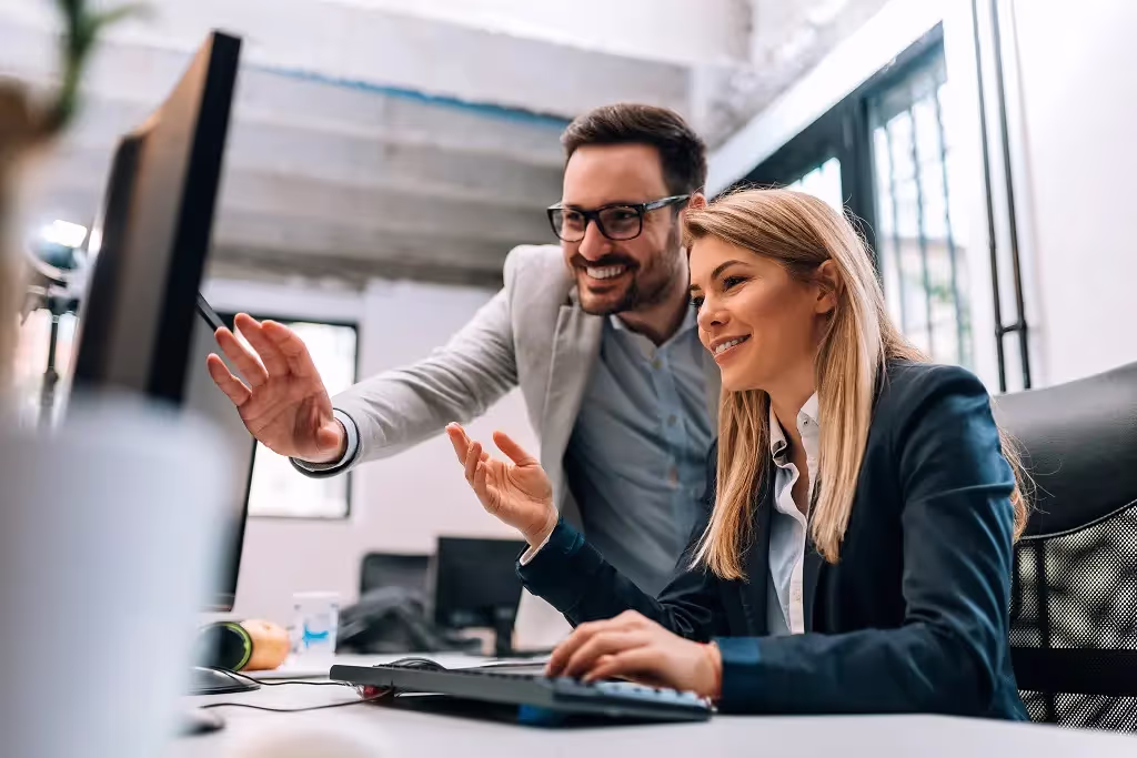 Two colleagues smiling and pointing at a computer screen in an office