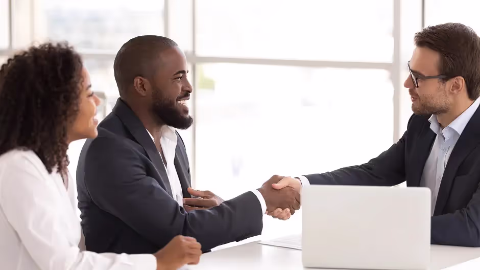Three professionals at a table, two shaking hands over a laptop
